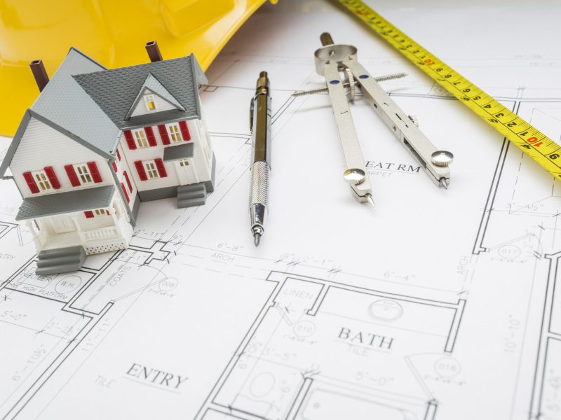 Model Home, Measuring Tape, Hard Hat, Pencil and Compass Resting on Custom House Plans.